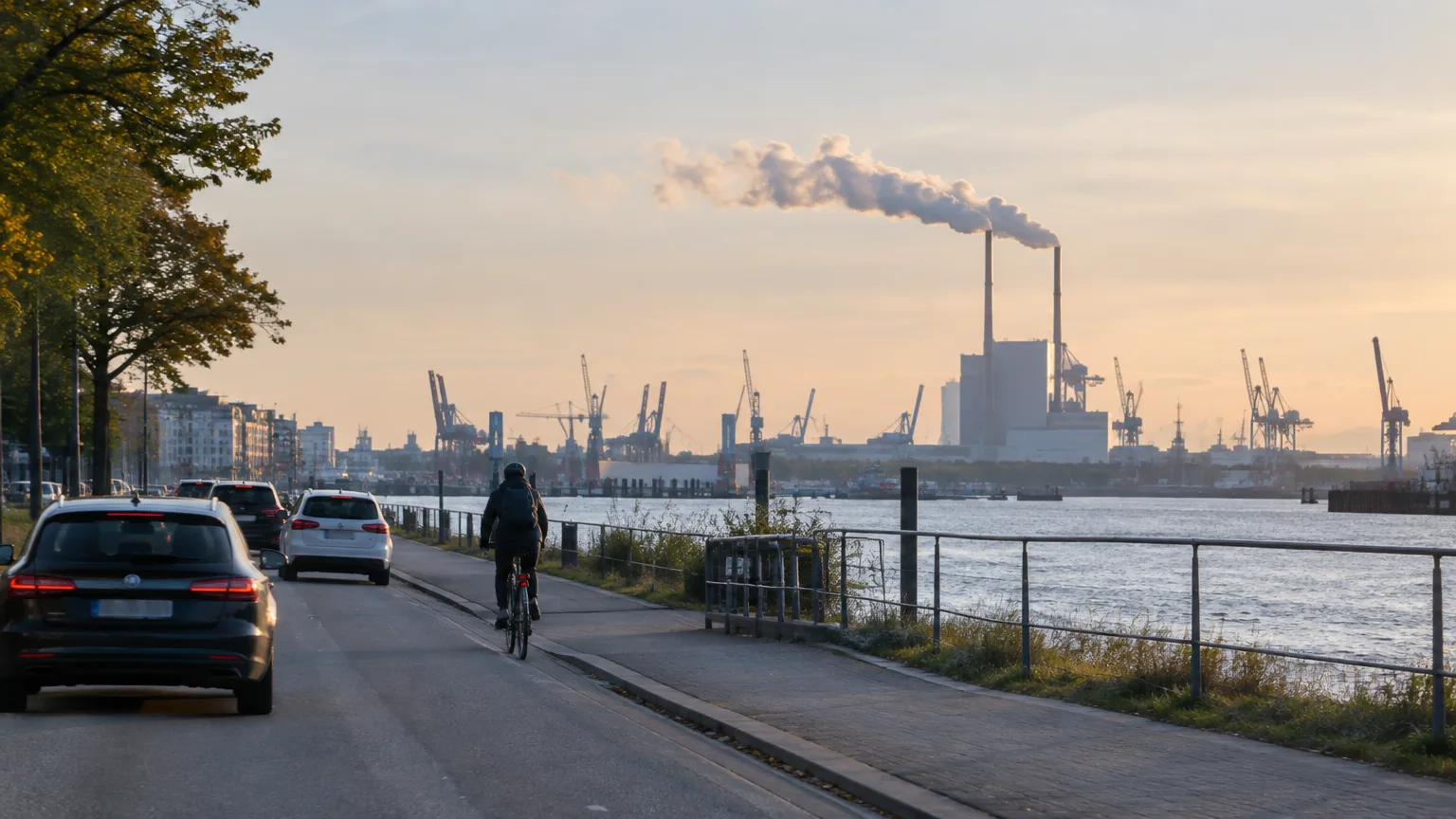 Eine Straße verläuft an einem Hafenbecken entlang; im Hintergrund stehen Kräne und rauchende Schornsteine.